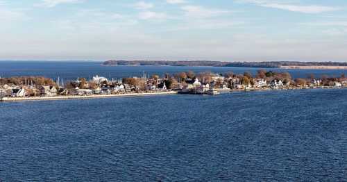 A scenic view of a coastal town along a calm blue bay, with trees and buildings lining the shore under a clear sky.