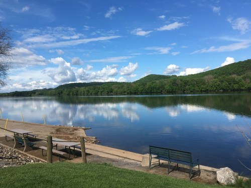 A serene lake scene with blue skies, fluffy clouds, and green hills reflected in the calm water, featuring benches by the shore.