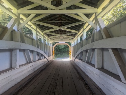 View of the interior of a covered bridge, featuring wooden beams and a pathway leading to bright light outside.
