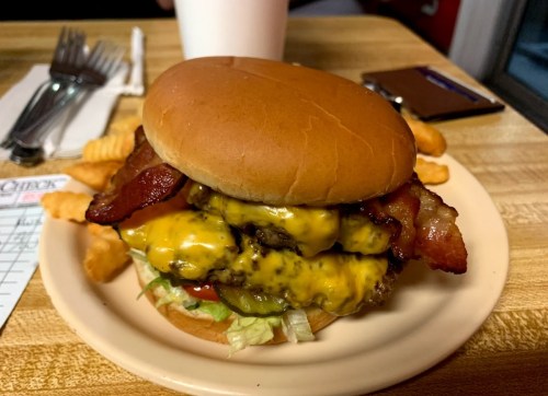 A large cheeseburger with bacon, lettuce, and condiments, served with crinkle-cut fries on a plate.