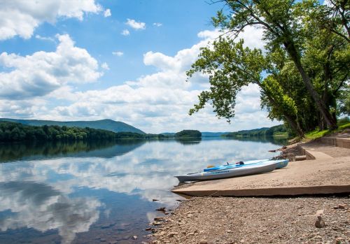 A calm river scene with a kayak on the shore, surrounded by trees and reflecting clouds in the water.