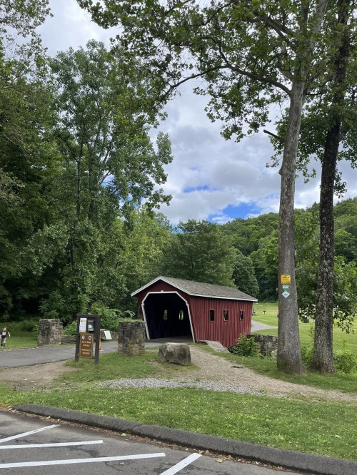 A red covered bridge surrounded by trees and a grassy area, with a cloudy sky in the background.