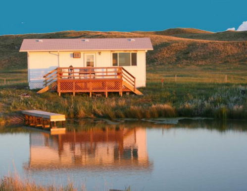 A small house with a wooden deck beside a calm pond, reflecting the structure and surrounding hills at sunset.