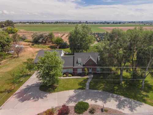 Aerial view of a house surrounded by trees and open fields, with a winding driveway and clear blue sky.