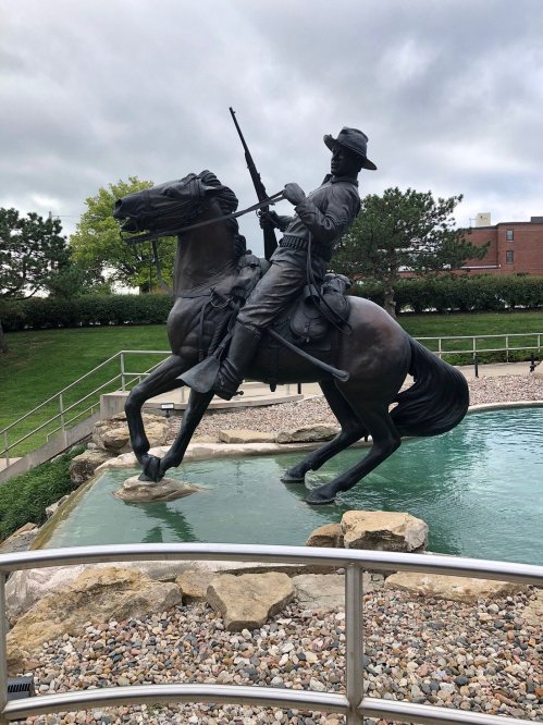 A bronze statue of a mounted soldier holding a rifle, rearing on a horse near a pond with rocks and greenery.