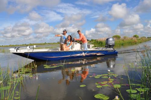 Three people on a blue boat navigating through a calm waterway with lily pads and lush greenery under a cloudy sky.
