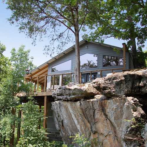 A modern house perched on a rocky ledge, surrounded by trees and greenery, with large windows and a wooden deck.