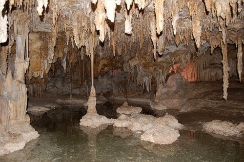 A dimly lit cave interior featuring stalactites, stalagmites, and a reflective pool of water on the ground.