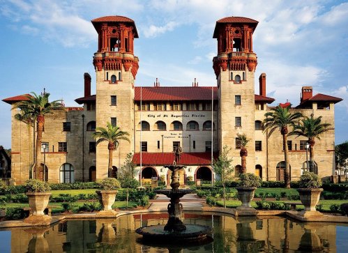 Historic building with red-tiled towers, surrounded by palm trees and a fountain in the foreground. Blue sky above.