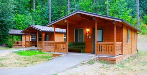 Two wooden cabins with porches, surrounded by green trees and grass, on a clear day.