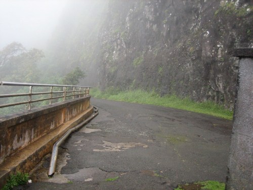 A misty road winding alongside a rocky cliff, with lush greenery and a railing on one side.