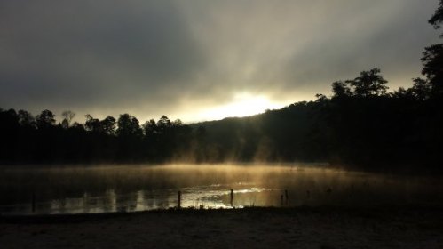 Misty lake at dawn with soft sunlight breaking through clouds and trees in the background.