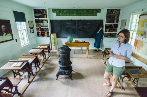 A woman stands in a vintage classroom with wooden desks, a chalkboard, and a globe, evoking a historical learning environment.