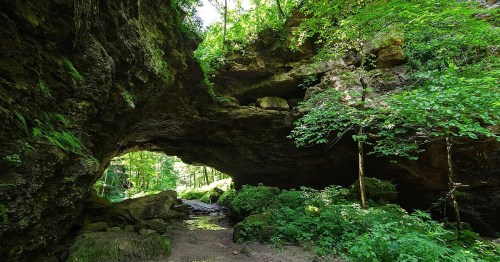 A natural rock arch surrounded by lush greenery and trees, with a small stream flowing beneath it.