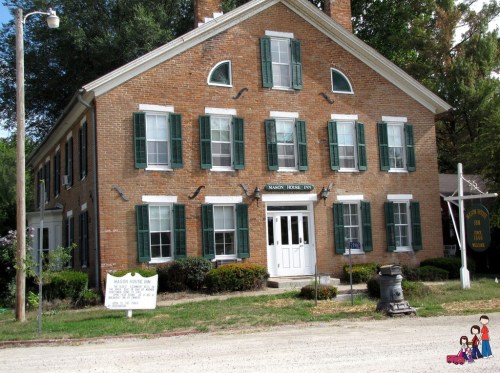 Historic brick inn with green shutters, white door, and sign, surrounded by greenery and gravel path.