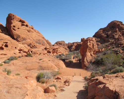 A scenic view of red rock formations and a sandy path in a desert landscape under a clear blue sky.