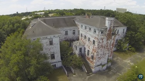 An aerial view of a dilapidated, overgrown building with peeling paint and a staircase leading to the entrance.