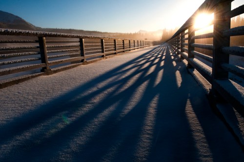 A snowy pathway with long shadows cast by a railing, illuminated by the sun rising in the background.