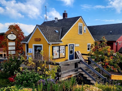 A bright yellow house with a sloped roof, surrounded by colorful foliage and flowers, features a welcoming sign.