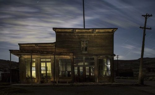 Abandoned wooden building at night, illuminated with a ghostly glow, under a cloudy sky with star trails.
