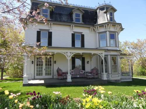 A charming Victorian-style house with a porch, surrounded by blooming flowers and trees on a sunny day.