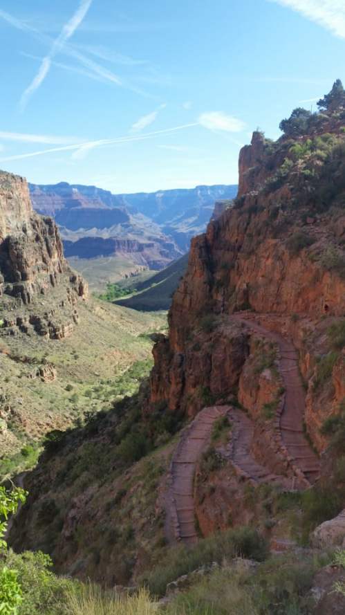 A winding trail leads through rocky terrain, surrounded by steep cliffs and distant mountains under a clear blue sky.