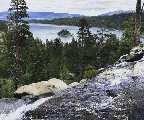 A scenic view of a lake surrounded by trees, with a waterfall in the foreground and mountains in the distance.