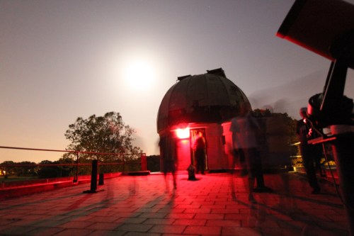 A dimly lit observatory at night, with a bright moon and blurred figures moving around the rooftop.