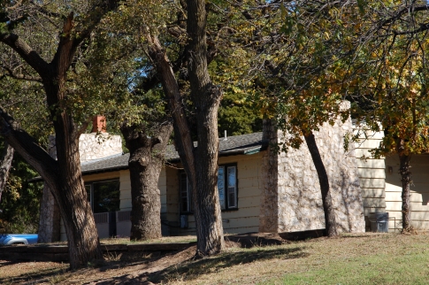 A rustic house surrounded by trees, featuring a stone chimney and a sloped yard.