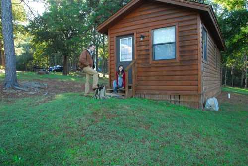 A wooden cabin in a wooded area, with a man standing and a woman sitting on the steps, accompanied by a small dog.