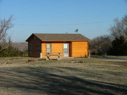 A small wooden cabin with a porch, surrounded by grass and trees under a clear blue sky.