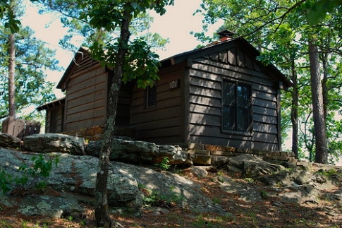 A rustic wooden cabin nestled among trees and rocks, with sunlight filtering through the leaves.