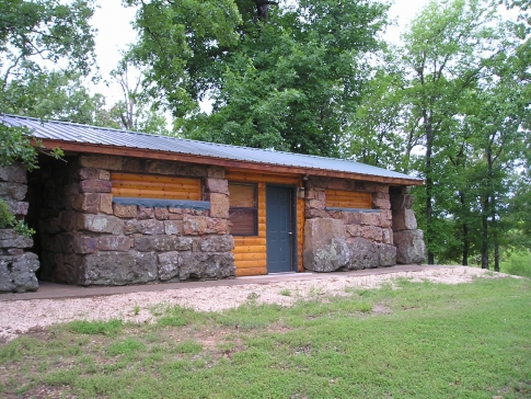 A rustic stone and wood cabin surrounded by trees, featuring a simple door and a gravel path.