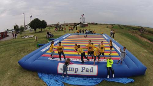 A colorful inflatable jump pad with children playing and jumping on it, set in a grassy outdoor area.