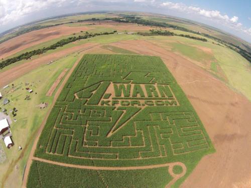 Aerial view of a corn maze featuring the logo "WARN" carved into the crops, surrounded by farmland and blue skies.