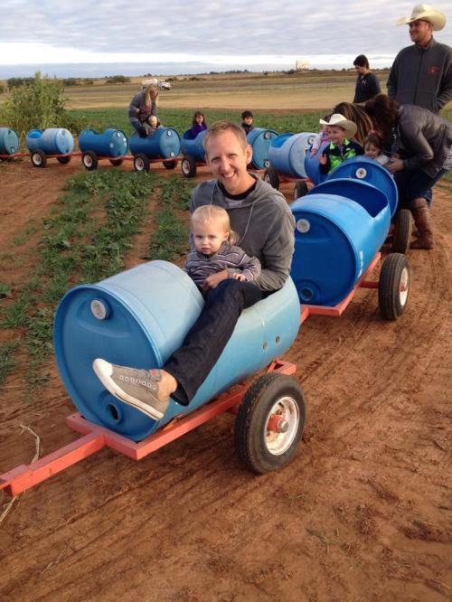A man and a child sit in a blue barrel on a cart, surrounded by people at a farm. Green plants are visible in the background.