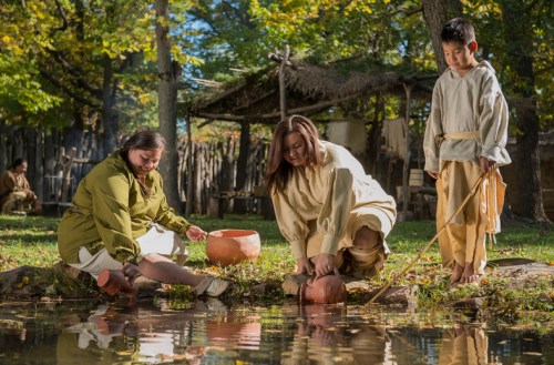 Three people in historical clothing gather by a pond, one filling a pot while others observe, surrounded by trees and a village.