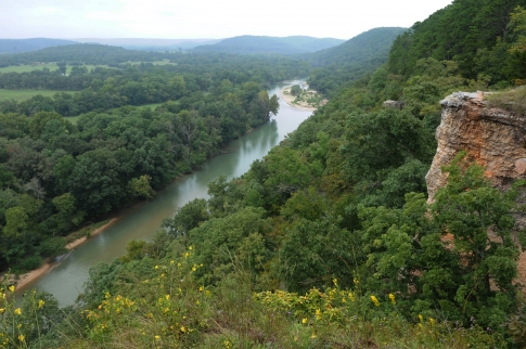 A scenic view of a winding river surrounded by lush green trees and rolling hills under a cloudy sky.