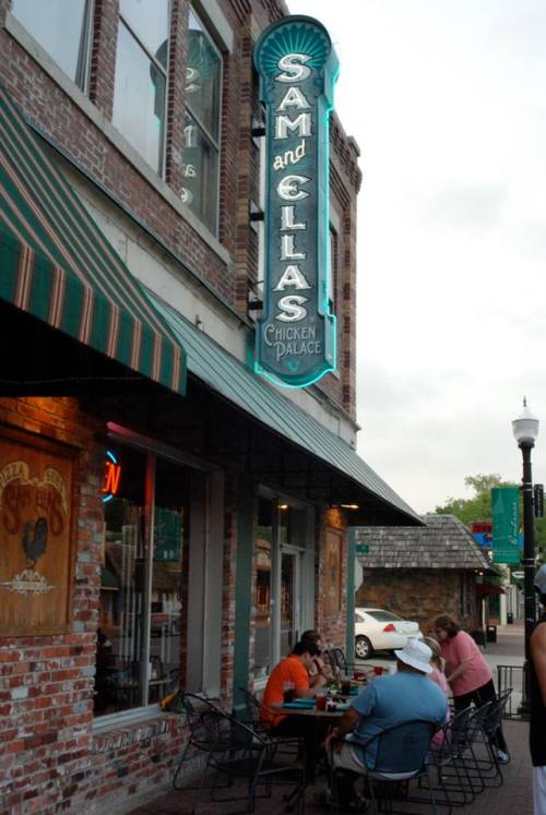 Exterior of Sam and Ella's Chicken Palace, featuring a neon sign and patrons dining outside on a brick sidewalk.