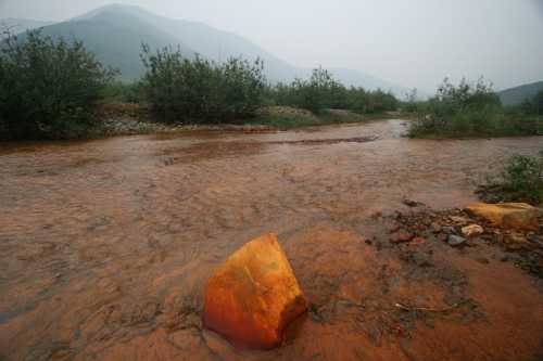 A rocky riverbed with orange-tinted water flowing through, surrounded by greenery and mountains in the background.