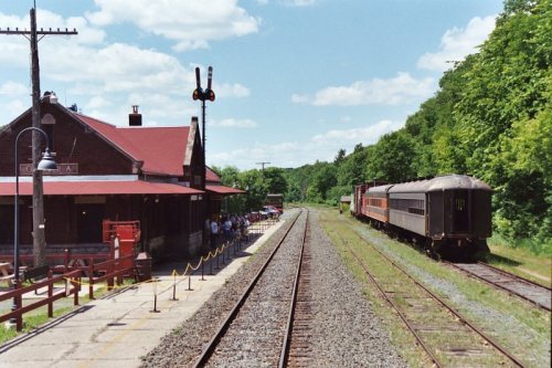 A train station with a red-roofed building, tracks, and a train on the right, surrounded by green trees and blue skies.
