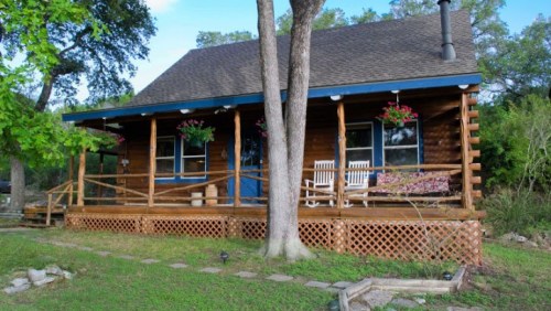 A charming wooden cabin with a porch, surrounded by greenery and hanging flower baskets.