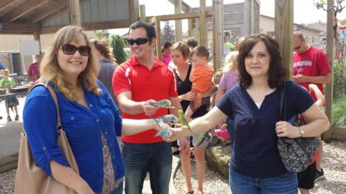 Two women and a man interact with children at a petting zoo, surrounded by families and playful animals.