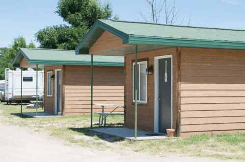 Two small wooden cabins with green roofs, set in a grassy area, with a table outside one of the cabins.