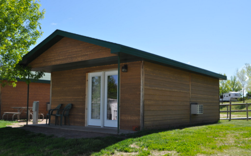 A small wooden cabin with a green roof, featuring a porch and large glass doors, surrounded by grass and trees.