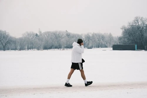 A person in a hoodie and shorts walks on a snowy path, surrounded by frosted trees in a winter landscape.