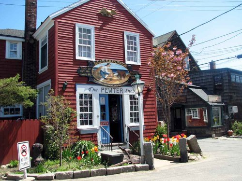 A charming red building with a sign reading "The Pewter Shop," surrounded by colorful flowers and a clear blue sky.