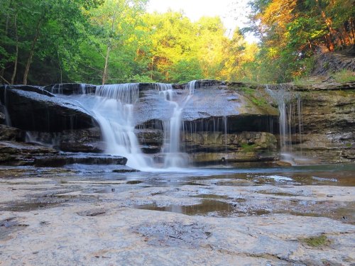 A serene waterfall cascading over rocky terrain, surrounded by lush green trees and warm sunlight filtering through.