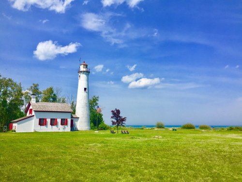 A white lighthouse beside a red-roofed building, surrounded by green grass and trees, with a blue sky and lake in the background.