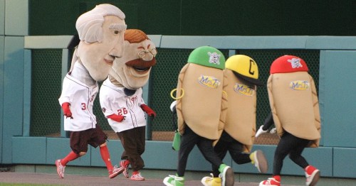 Four mascot characters, including two large heads and two taco costumes, race on a baseball field.